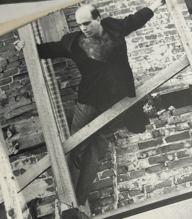 Michael A. Carson posed against a brick wall with wooden beams in an Ausdruckstanz Dance Theatre publicity photo